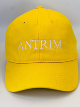 Load image into Gallery viewer, Yellow  baseball cap with &#39;ANTRIM&#39; embroidered in white thread. Adjustable back strap with metal clasp.
