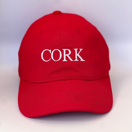 Red baseball cap with 'Cork' embroidered in white thread. Adjustable back strap with metal clasp.