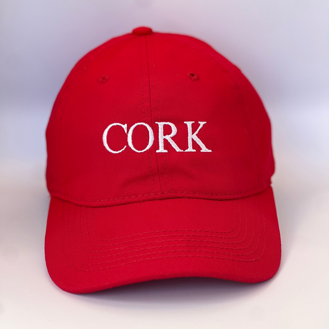 Red baseball cap with 'Cork' embroidered in white thread. Adjustable back strap with metal clasp.